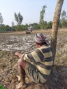 A farmer taking rest under a tree observing his son pulling tractor in the muddy field.