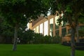 Sunlight reflects on the windows of a multi-storey building among thickets of trees over a lawn with birds.