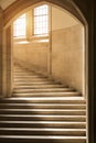 Sunlight shining through windows onto a classic, gothic style stone stairway curving upward through an archway