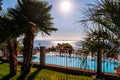 Sunny outdoor pool behind the fence with crystal water, parasols and deck chairs with Garda lake and Sirmione city on the