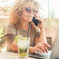 Sunny portrait of cheerful busy modern business woman busy in phone call and writing on laptop sitting at summer cafe bar with Royalty Free Stock Photo