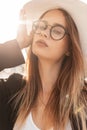 Sunny portrait of a pretty young fashionable woman in glasses and a white hat with a black jacket and t-shirt at sunset. Beachwear Royalty Free Stock Photo