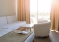 Sunny room in the hotel in light colors with a bed, white table, sofa and armchair. New white disposable slippers in a