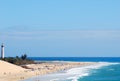 A sunny sand beach being hit gently by foamy wave crests, with a lighthouse standing on a pouch of tropical trees