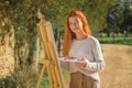 Sunny shot of a redhead female painting on a painting board with palettes in a park