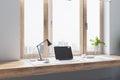 Sunny stylish work place with wooden table top near big window with city view, modern laptop and plant in glass vase