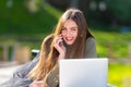 On a sunny and windy day young smiling woman lying on bench in park and working on laptop and talking on a smartphone