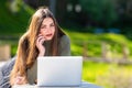 On a sunny and windy day young smiling woman lying on bench in park and working on laptop and talking on a smartphone