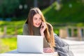 On a sunny and windy day young smiling woman lying on bench in park and working on laptop and talking on a smartphone