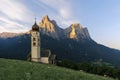 Sunrise landscapes of Church St. Valentin on grassy hilltop with view of rugged peaks of Mountain Schlern with alpenglow in backgr