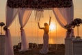 Sunset. bride silhouette. Wedding ceremony arch with flower arrangement and white curtain on cliff above sea, outdoor photo.