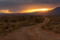 Sunset light glows orange over a landscape with a dirt road leading into the desert with the sun going down behind clouds Royalty Free Stock Photo