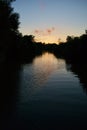 Sunset on a narrow river: silhouetted, shadowed trees, clouds, golden sky reflected in water