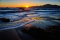 Sunset over distant mountains with blue waves crashing into sandy beach Royalty Free Stock Photo