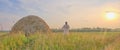 At sunset an senior Muslim man in an embroidered skullcap and white traditional Clothes mows hand-scythe grass in a hayfield