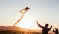 Sunset sun backlit silhouettes of a daughter with father while they trying to launch a colorful kite in the mountain fields. Warm Royalty Free Stock Photo