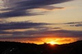 Sunset time over silhouettes of hills, houses roofs and forest. Dark purple long lines clouds in the sky. Royalty Free Stock Photo