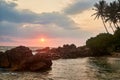 Sunset at tropical beach with golden sky reflecting on calm water. Palm trees silhouette, rocky shoreline at dusk Royalty Free Stock Photo