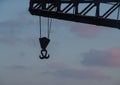 Sunset view of a metal double hook hanging with ropes from a tall crane in front of a cloudy blue pink sky