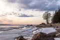 Sunset winter lake shore landscape with stones, snow and ice, bare trees, cloudy sky. Ladoga lake Royalty Free Stock Photo