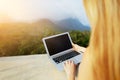 Sunshine photo of blonde girl using laptop, mountains in background, Thailand.