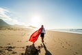 Superhero child walking on the sandy, sunset beach, wearing red cape. Back view. Full length. Summer vacation concept