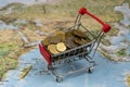 A supermarket cart full of coins. On the background is a map of the world. It symbolizes finance, investment, and the