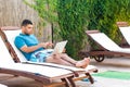 Suprised bearded young adult freelancer man in blue t-shirt and shorts lying on deck chair on poolside, pointing finger and