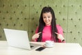 Suprised young girl with black dreadlocks hairstyle in pink blouse sitting in cafe and having new proposal for work on laptop,
