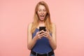 Suprised young pretty long haired redhead woman in blue shirt looking excitedly on screen of her smartphone while reading Royalty Free Stock Photo