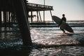 Surfboarder carrying a surfboard in the wavy ocean near the wooden pier during sunset Royalty Free Stock Photo