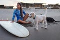 Surfer Girl. Surfing Woman On Beach Near Surfboard And Dog. Smiling Female In Blue Wetsuit Sitting On Sand. Royalty Free Stock Photo