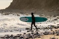 Surfer walking into ocean with board as waves crash on a rocky shore. Royalty Free Stock Photo