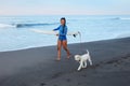 Surfing Girl. Beautiful Woman Walking With Dog On Ocean Beach. Brunette In Blue Wetsuit Carrying Surfboard. Royalty Free Stock Photo