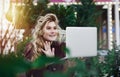 Thoughtful young woman in glasses using a computer, sitting on a bench in a city park. The concept of time is online and work in