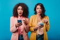 Surprised african girl in pink jacket holding phone. Two brunette women holding smartphones