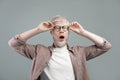 Surprised albino blond man touching glasses with open mouth, guy posing over grey studio background