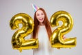 Surprised and amazed radhaired ginger woman with birthday cap horn holding ballons 22 years anniversary numeral in Royalty Free Stock Photo