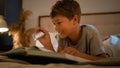 Surprised amazed smiling funny Caucasian child kid boy schoolboy reading interesting book evening in bedroom using Royalty Free Stock Photo