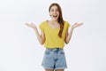 Surprised and amused charismatic attractive woman in yellow t-shirt shrugging with hands spread near shoulders open