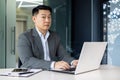 Surprised asian man looking at camera, mature adult boss businessman working inside office with laptop while sitting at Royalty Free Stock Photo