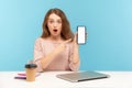 Surprised astonished woman, office employee, sitting at workplace and pointing cellphone mock up display, copy space Royalty Free Stock Photo