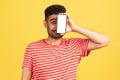 Surprised bearded man in red striped t-shirt hiding face behind smartphone with empty display, having fun, trying new application