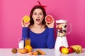 Surprised beautiful woman holds slice of orange and grapefruit, has shocked expression, opens mouth from amazement, surrounded