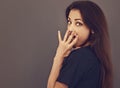 Surprised beautiful woman looking with big eyes, opened mouth and covering mouth the hand in black shirt on grey background. Royalty Free Stock Photo