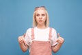 Surprised blond girl in cute clothes stands on a blue background, showing fingers down and looking in camera.Teenage girl with a Royalty Free Stock Photo