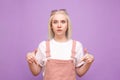 Surprised blond girl in cute clothes stands on a purple background, showing fingers down and looking in camera.Teenage girl with a Royalty Free Stock Photo