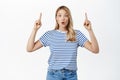 Surprised blond girl in striped t-shirt see big news, points fingers up, reacts amazed and impressed, fascinated by Royalty Free Stock Photo