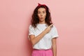 Surprised brunette woman, shows finger to the copy space isolated over pink background, wears casual white t-shirt and Royalty Free Stock Photo