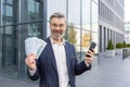 Surprised businessman holding pile of money and phone in hands, happy with winning lottery, promotion at work, looking Royalty Free Stock Photo